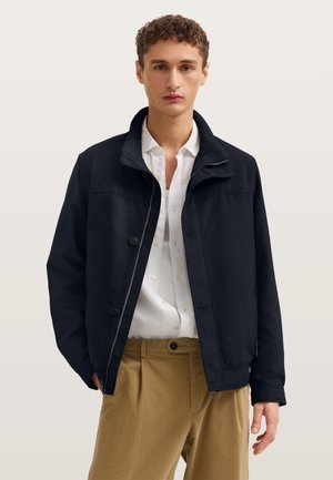 Junger Mann, der eine schwarze Jacke über einem weißen Hemd mit Knöpfen und beigefarbenen Hosen trägt, vor einem schlichten hellen Hintergrund stehend.