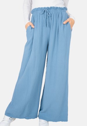 Person trägt lockere, hellblaue, weite Hose mit Kordelzug in der Taille und Taschen, kombiniert mit einem weißen Langarmoberteil und weißen Sneakers.