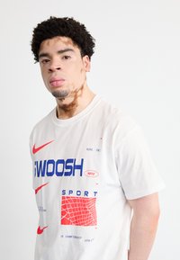 Balts kokvilnas t-krekls ar zilu un sarkanu grafisko apdruku, kurā ietilpst "SWOOSH" un Nike logo, kā arī ar fakturētu sarkanu režģa rakstu.