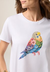 Biała koszulka z kolorowym wzorem ptaka, utworzonym z kwiatowych wzorów i kamieni szlachetnych, z klasycznym okrągłym dekoltem i krótkimi rękawami.