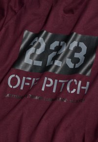 Mörkröd tyg med fet grå text som visar "223 OFF PITCH" med koordinater nedanför. Mjuk textur med en något skrynklig yta.