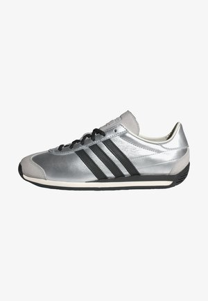 Zapatilla deportiva plateada con superficie lisa de cuero, diseño de tres rayas negras y cordones negros. Presenta una puntera redondeada y suela de goma.