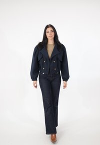 Marineblå cropped jakke med stor krave, dobbeltradede knapper og puffede ærmer, kombineret med mørke udvidede jeans og brune ankelstøvler.