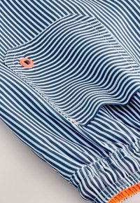 Triibuline kangas tumesinise ja valgega, elastse vöökohaga ja oranžide detailidega. Omab plaastri-taskut nähtava õmbluse ja metallkraega.