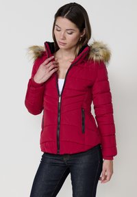 Raudona pūkinė striukė su dirbtinio kailio kapšeliu, užtrauktuko priekyje, siuvinėta tekstūra ir šoninėmis užtrauktuko kišenėmis. Dėvima ant baltos palaidinės ir tamsių džinsų.