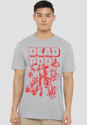 Junger Mann trägt ein graues T-Shirt mit roten Deadpool-Figuren und Text auf der Vorderseite vor einfachem Hintergrund.