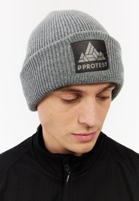 Grijze gebreide beanie met een geribbelde textuur. Heeft een zwart label met een geometrisch ontwerp en het woord "PROTEST". Geschikt voor koud weer.