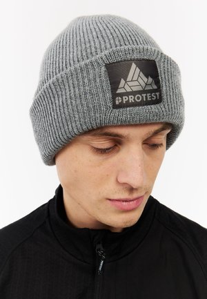 Gray knit beanie with a ribbed texture. Features a black patch with a geometric design and the word “PROTEST.” Suitable for cold weather.