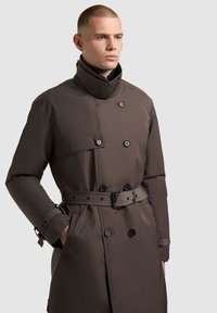 Brown trench coat featuring double-breasted buttons, a belted waist, and a high collar. Made from a smooth, weather-resistant material.