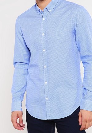 Chemise d'un bleu clair avec un motif texturé, à manches longues et ourlet arrondi. Présente des boutons blancs et un col.
