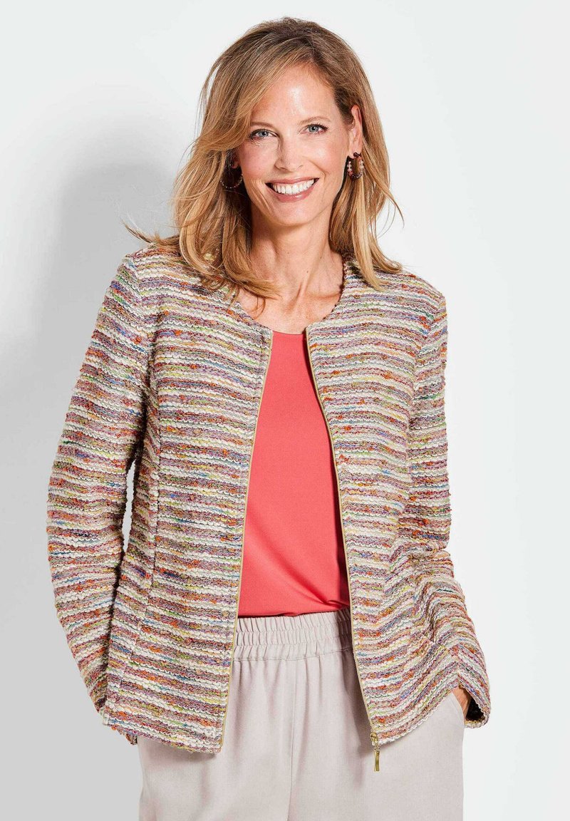 Giacca con zip frontale in tessuto a righe multicolori con zip dorata, abbinata a una maglietta corallo e pantaloni beige chiaro. Vestibilità casual, tessuto leggero.