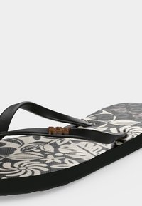Σανδάλι flip-flop με μαύρα λουριά και σόλα με ασπρόμαυρο floral μοτίβο, με μικρή καφέ λεπτομέρεια λογότυπου στο λουράκι.