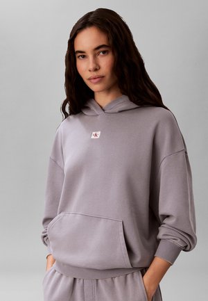 Frau trägt einen lockeren hellvioletten Hoodie mit kleinem Logo auf der Brust und passende Hose, steht mit Händen in den Taschen.