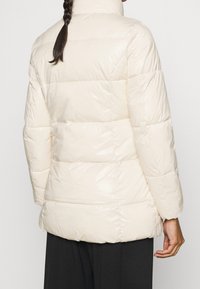 Donna che indossa un giubbotto imbottito a trapunta di colore crema con maniche lunghe e pantaloni neri, vista da dietro su uno sfondo semplice.