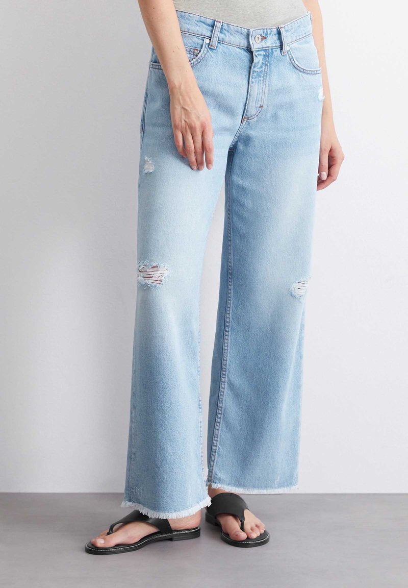 Jasnoniebieskie dżinsy w stylu wide-leg, wykonane z denimu, z przetartymi miejscami, postrzępionymi brzegami i klasycznym pięci pockets. Noszone z czarnymi klapkami.