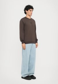Maglione marrone a maniche lunghe con scollo rotondo e logo, abbinato a jeans a gamba larga blu chiaro e scarpe nere, su uno sfondo bianco.