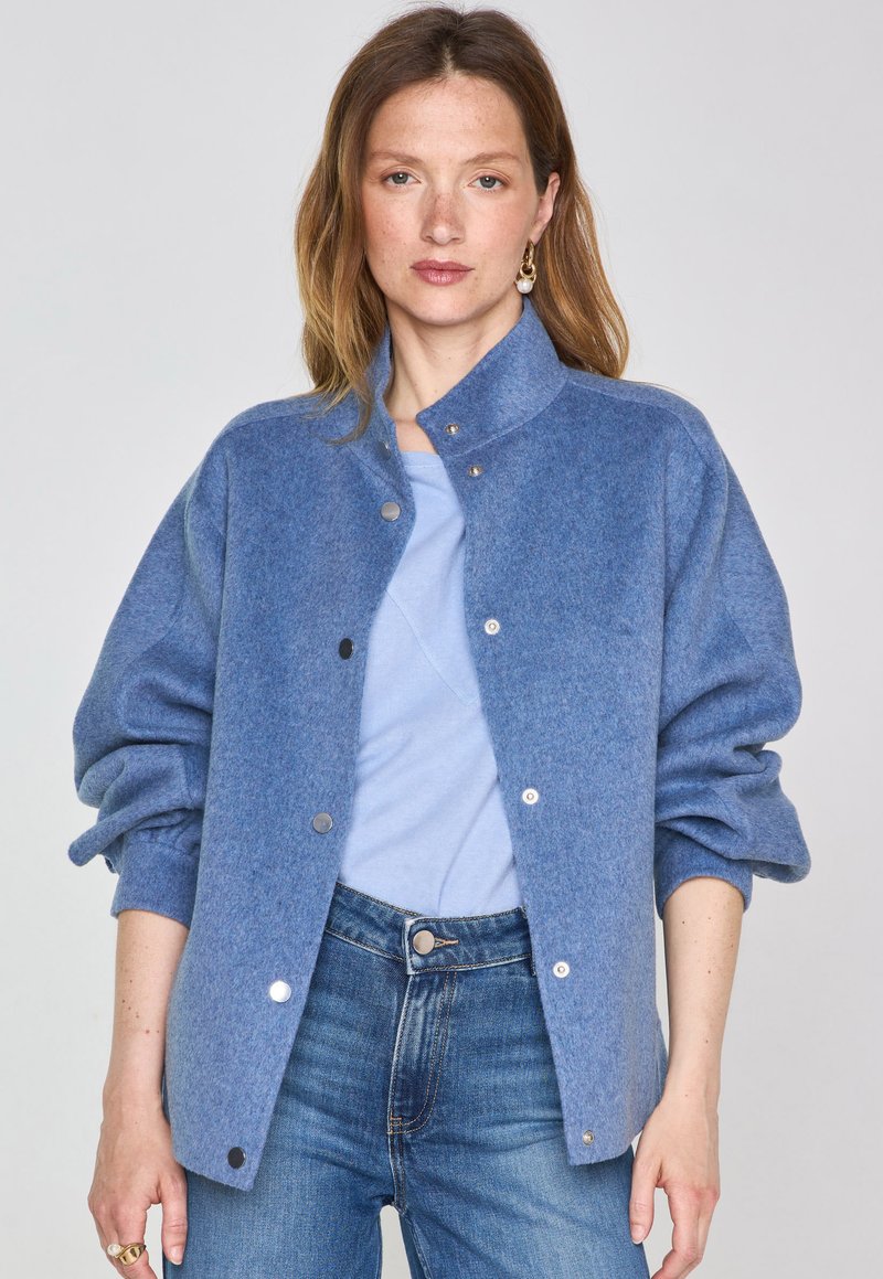 Giacca di lana blu con colletto alto, chiusura a bottoni a pressione e maniche oversize, abbinata a una maglietta azzurra e jeans in denim.