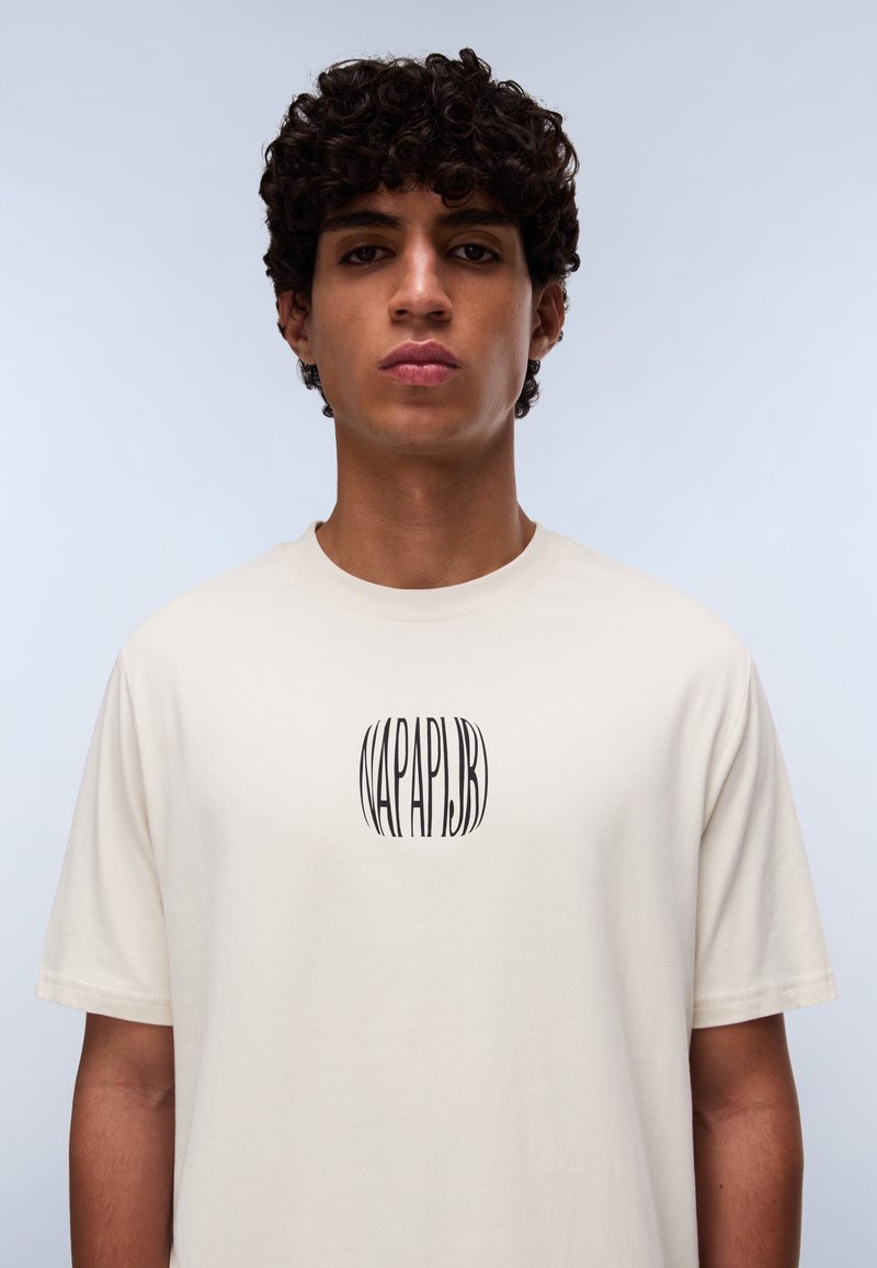 Cremefarbendes Baumwoll-T-Shirt mit kurzen Ärmeln, das ein zentrales schwarzes Logo mit der Aufschrift "NAPAPIJR" auf der Brust zeigt. Einfaches Design und weiche Textur.