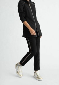 Czarna kurtka zapinana na zamek z srebrnymi akcentami, w połączeniu z czarnymi spodniami z metalicznymi paskami po bokach oraz białymi trampkami ze złotymi detalami.