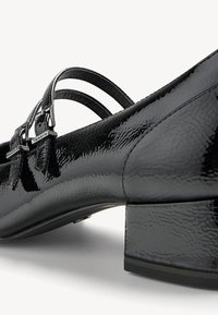 Chaussure noire brillante à talon bas avec doubles fines lanières et petites boucles argentées, montrant une surface en cuir verni texturée.