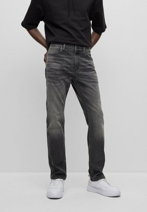 Osoba w czarnej bluzie z kapturem, wyblakłych czarnych dżinsach i białych trampkach, stojąca z rękami za plecami na jednolitym szarym tle.