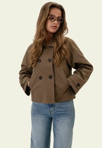 Femme aux longs cheveux ondulés portant des lunettes écaille, une veste marron croisée avec des boutons, et un jean bleu clair, mains dans les poches.