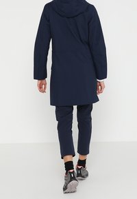 Mörkblå vattentät jacka med huva, bakvy. Har en medellång design och bärs med matchande byxor och grå vandringsskor.