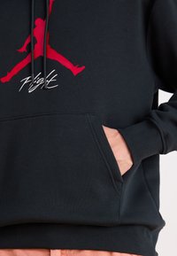 Sweatshirt negru cu un logo grafic roșu și text alb "Flight". Are un buzunar frontal și glugă cu șnur. Material moale, texturat.