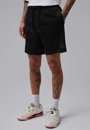 Beine mit Tattoos, die schwarze Netzshorts, weiße Socken und weiße und rote Sportschuhe auf einfarbigem Hintergrund tragen.