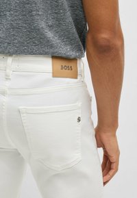 Białe dżinsy z denimu z skórzaną metką BOSS na pasku oraz srebrnymi akcentami na tylnych kieszeniach. Posiadają standardowy wzór kieszeni.