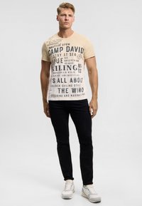 Beige short-sleeve t-shirt with large black text and nautical-themed graphics, paired with fitted black jeans and white athletic sneakers.