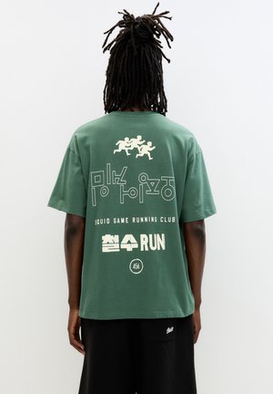 Groene katoenen t-shirt met een groot wit gedrukt ontwerp met stijlvol tekst, waaronder "Squid Game Running Club" en drie hardlopende figuren.