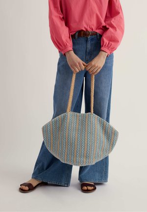 Persona con blusa rosa y jeans azules sosteniendo un bolso tote tejido a rayas con asas de cuerda, usando sandalias marrones.