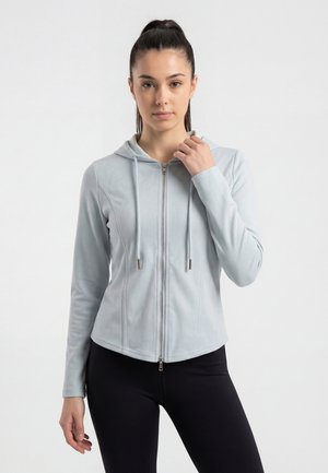 Jeune femme aux cheveux attachés porte un sweat à capuche gris clair zippé et des leggings noirs, posant devant un fond blanc uni.