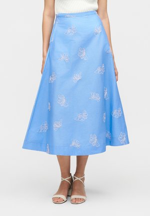 Kobieta w jasnoniebieskiej spódnicy midi z białym kwiatowym haftem i białych sandałach na obcasie z paskami, stojąca na jednolitym tle.