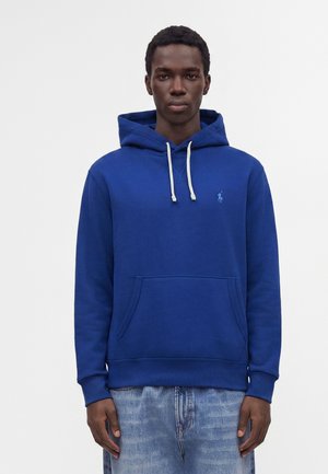 Jeune homme portant un sweat à capuche bleu avec des cordons blancs et un jean bleu clair, debout devant un fond blanc uni.