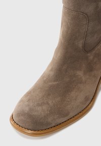 Braune Wildleder-Ankle-Boots mit glatter Textur, abgerundeter Zehenpartie und genähten, hellbraunen Sohlenakzenten. Minimalistisches Design ohne sichtbare Hardware-Elemente.