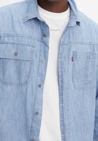 Camicia in chambray azzurro chiaro con due tasche frontali, chiusura con bottoni e bottoni in tono argento, indossata sopra una maglietta bianca.