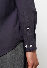 Camicia a maniche lunghe di un marrone scuro, con polsini a punta, caratterizzata da bottoni bianchi e una superficie liscia. Progettata con un orlo dritto e un design minimalista.