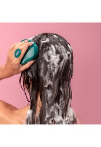 Rund teal shampooborste i användning på blött, lödderigt hår, med vita bubblor och en rosa bakgrund. Håret ser mörkt och vågigt ut.