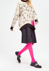 Pull beige oversize avec des motifs de chats noirs, associé à une jupe noire et des collants rose vif, complété par des chaussures noires à lacets brillantes.
