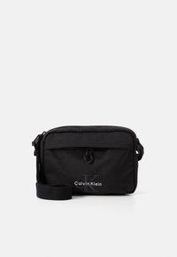Zwarte nylon schoudertas met ritsluiting, verstelbare schouderband en reliëf "Calvin Klein" logo aan de voorkant. Compact rechthoekig ontwerp.
