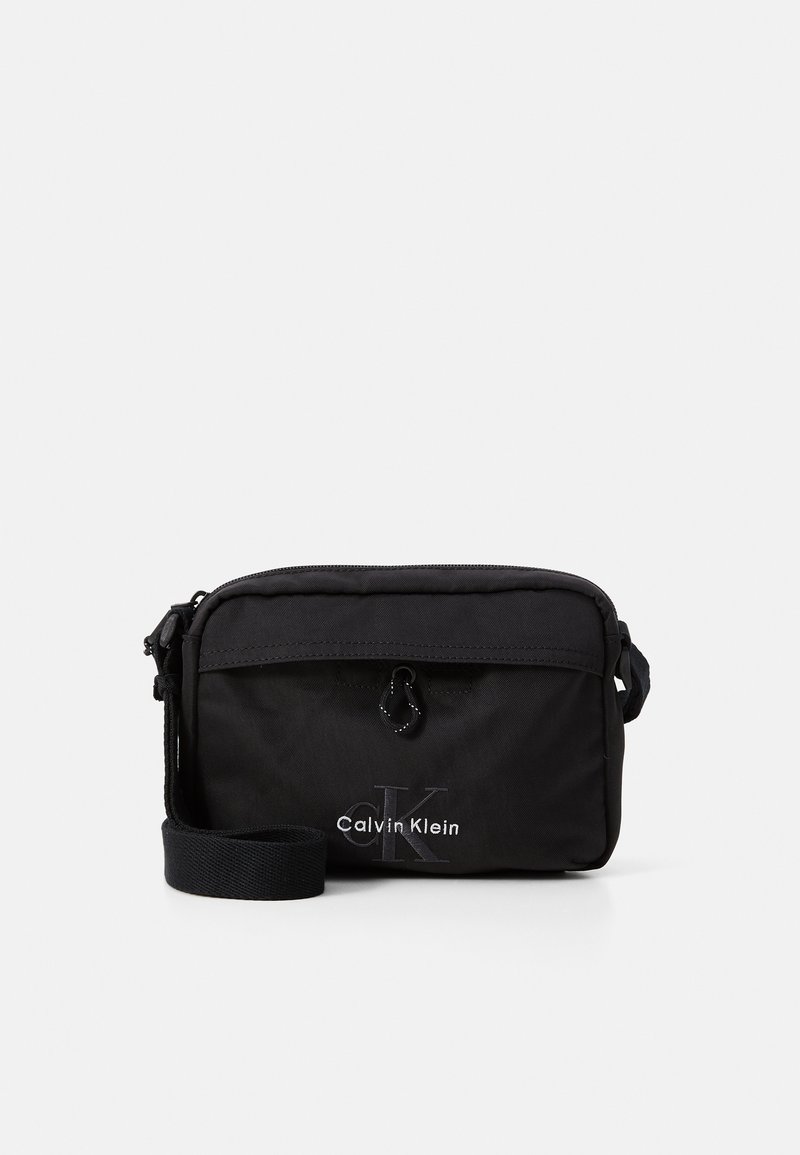 Zwarte nylon schoudertas met ritsluiting, verstelbare schouderband en reliëf "Calvin Klein" logo aan de voorkant. Compact rechthoekig ontwerp.