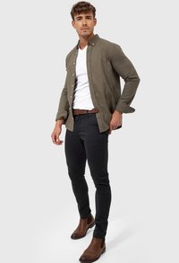 Camisa de botones color verde oliva sobre una camiseta blanca de cuello en V, combinada con pantalones negros ajustados y botas Chelsea marrones. Textura de tela suave.