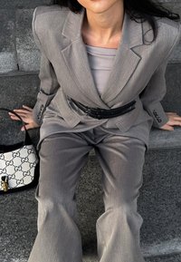 Conjunto de dos piezas gris con un acabado texturizado, que incluye una chaqueta ajustada y pantalones acampanados. Cinturón negro, blusa clara y un bolso blanco con patrón negro.