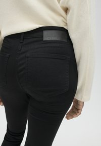 Persoon draagt zwarte high waist jeans met achterzakken en een crèmekleurige, gestructureerde top met lange mouwen, afgebeeld van de taille naar beneden.