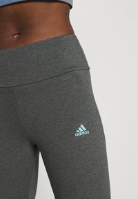 Grå träningstights med bred midja och en präglad ljusblå Adidas-logotyp. Tyget har en slät, strukturerad yta.