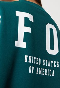 Tyrkysová mikina vyrobená z směsi bavlny, s velkými bílými písmeny "F" a "O" a textem "UNITED STATES OF AMERICA" v tučném písmu.