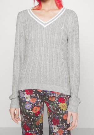 Osoba ubrana w jasnoszary sweter z dekoltem w serek o splocie warkoczowym oraz wielokolorowe spodnie w kwiaty, stojąca na jednolitym tle.