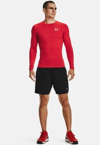 Rød langærmet sportshemse, sorte shorts og røde løbesko. Hemsen har en tætsiddende design, glat tekstur og logo på brystet.
